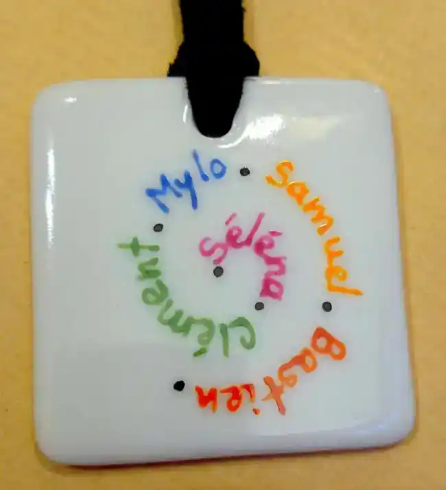 Pendentif carré en porcelaine personnalisé par des prénoms de différentes couleurs en spirale.